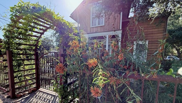 Een houten huis met een veranda, omringd door een tuin met bloeiende planten en een metalen hek.