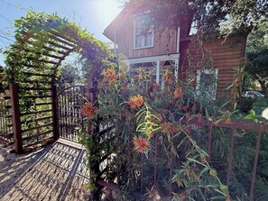 Ein Holzhaus mit einer Veranda, umgeben von einem Garten mit blühenden Pflanzen und einem Metallzaun.