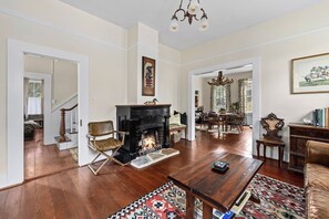Uma sala de estar ampla com lareira, piso de madeira e uma área de jantar visível através de uma porta aberta.