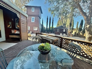 Una terraza de madera con una mesa y sillas de vidrio, un pequeño jardín con cercado de madera, y un edificio marrón con una ventana.