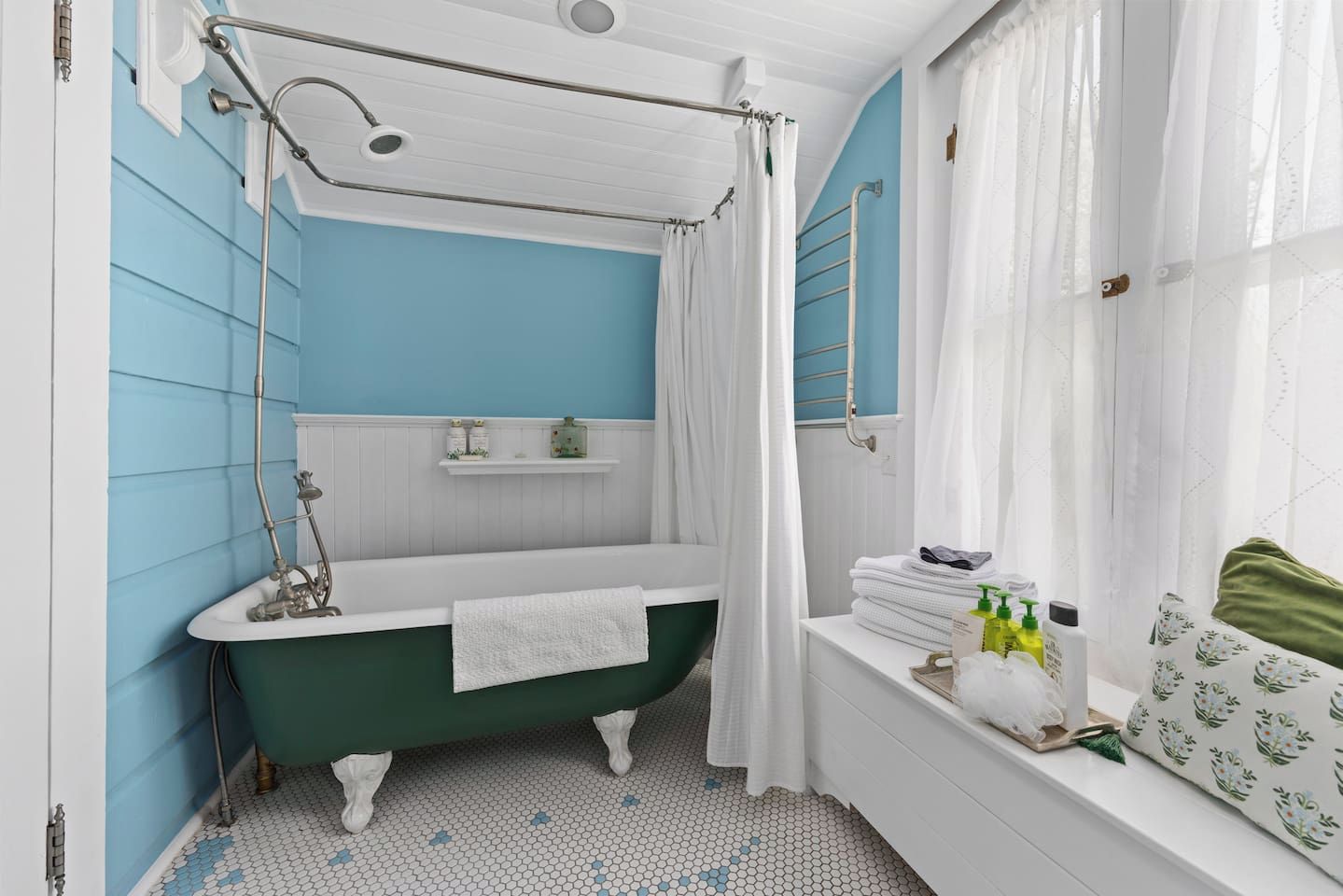 A bathroom with a freestanding bathtub, a showerhead, a window with curtains, and a shelf with toiletries.
