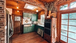 A rustic kitchen with wooden walls, a brick fireplace, and a large window. - 1882 Heritage House ~ Porches, Gardens & Bali Bed (Los Alamos)