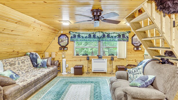 Een gezellig cabin interieur met houten plafond, plafondventilator, bank, stoel, raam met uitzicht op bomen, en een trap.