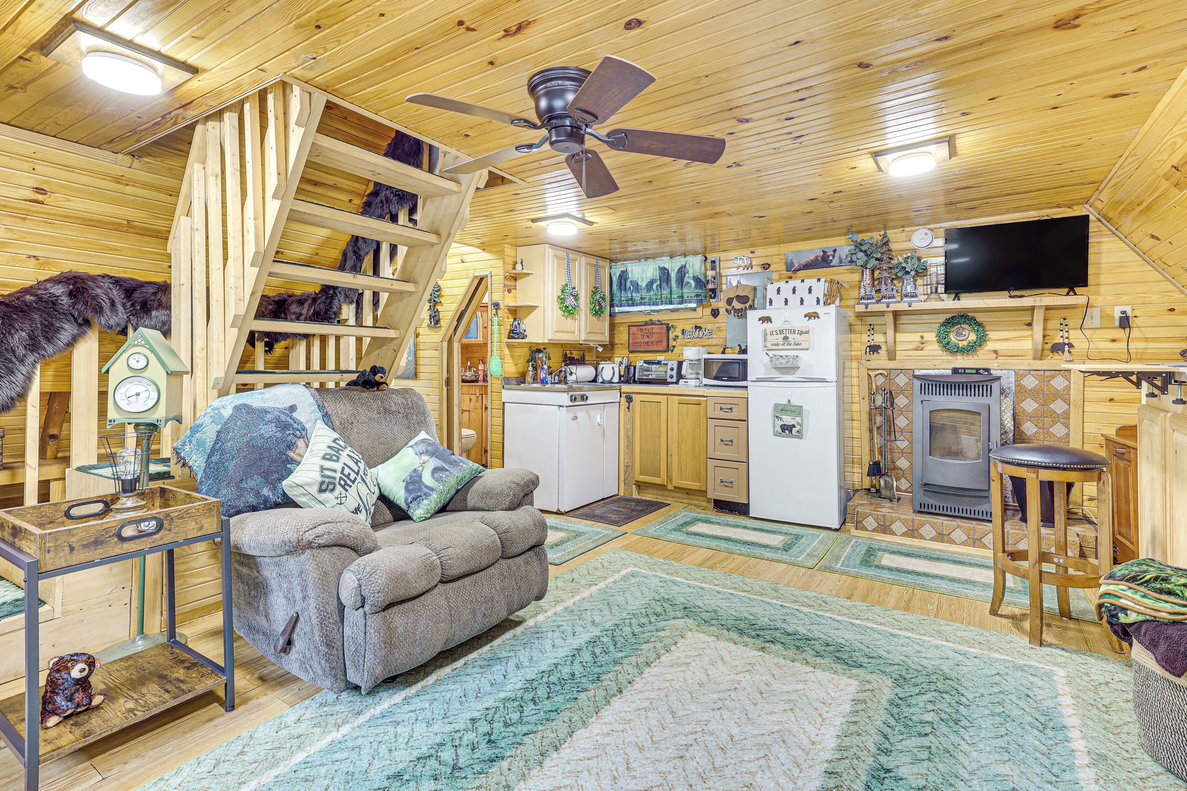 A cozy cabin interior with a sofa, wooden walls, a ceiling fan, a kitchen area, and a fireplace.