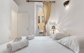 A hotel room with a bed, two rolled towels, a nightstand, a lamp, and a view of the outside.