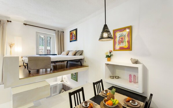 A modern, well-lit room with a bed, a dining table with chairs, and a small kitchen area.