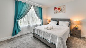 Ein ordentlich bezogenes Bett mit weißer Decke und Kissen, ein Nachttisch mit Lampe und ein Fenster mit türkisblauen Vorhängen.