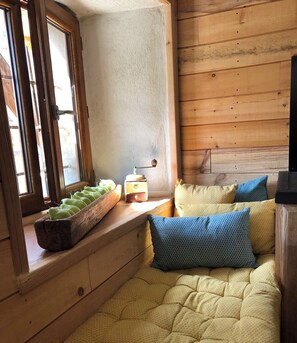 Un coin lecture confortable avec un banc en bois, une petite table sur laquelle repose un livre et une fenêtre donnant sur les montagnes.