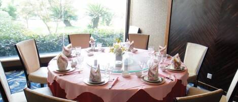 A round dining table with a pink tablecloth, surrounded by four chairs, set with white plates, glasses, and a vase of flowers.