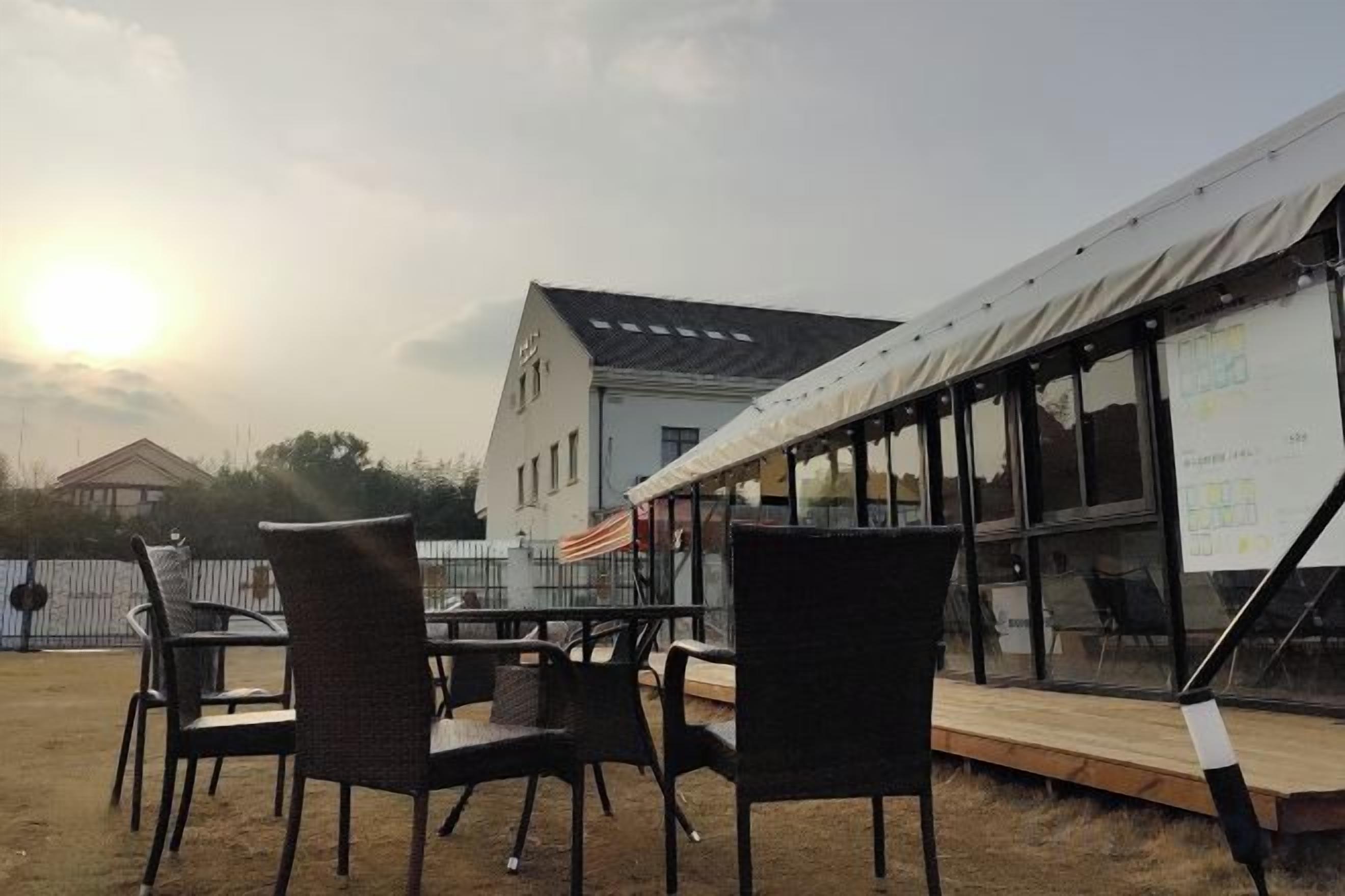 An outdoor seating area with wicker chairs and a wooden table, a tent structure, and a building with large windows in the background.