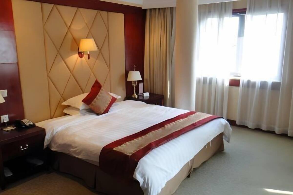 Habitación de hotel con una cama grande, cabecero con lámpara, mesita de noche con lámpara y ventana con cortinas.
