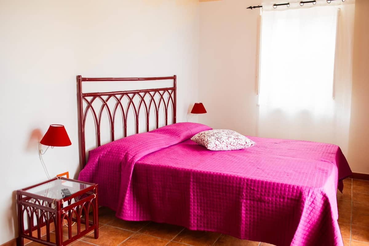 A bedroom with a wooden bed frame, a glass side table, a red lamp, and a window with a white curtain.
