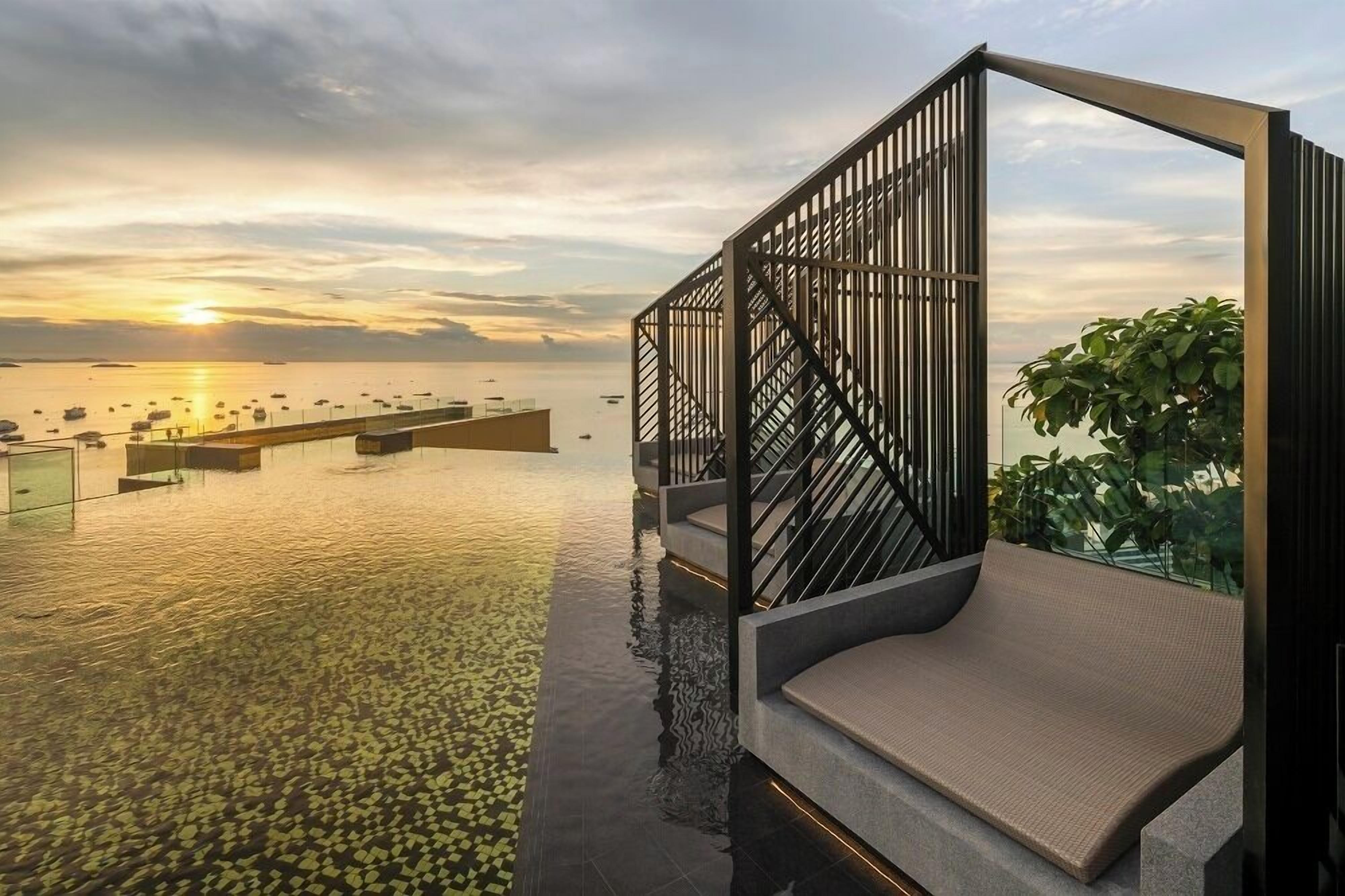 An infinity pool with a view of the sunset over the water, a modern outdoor seating area, and a glass railing.
