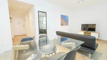 Un salón moderno con una mesa de vidrio, dos sillones grises, un sofá negro, un televisor sobre un mueble de madera y un cuadro en la pared.