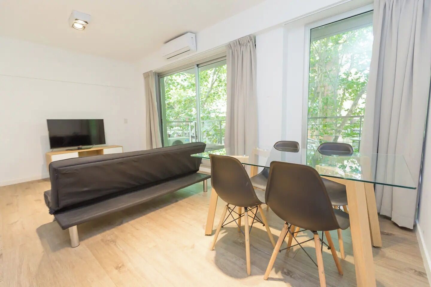 Una sala de estar moderna con un sofá negro, una mesa de comedor de vidrio con cuatro sillas, un televisor sobre un mueble de madera y ventanas grandes con cortinas.