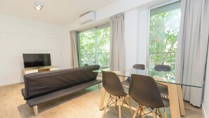 A modern living room with a black sofa, a glass dining table with four chairs, a TV on a wooden stand, and large windows with curtains. - Apartamento de 2 Ambientes en Belgrano (Buenos Aires)