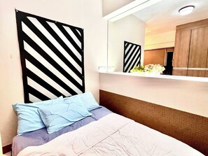 A bedroom with a bed, pillows, a striped partition, a mirror, and a small shelf with flowers. - Greenfield Ortigas CBD Residences (Mandaluyong)