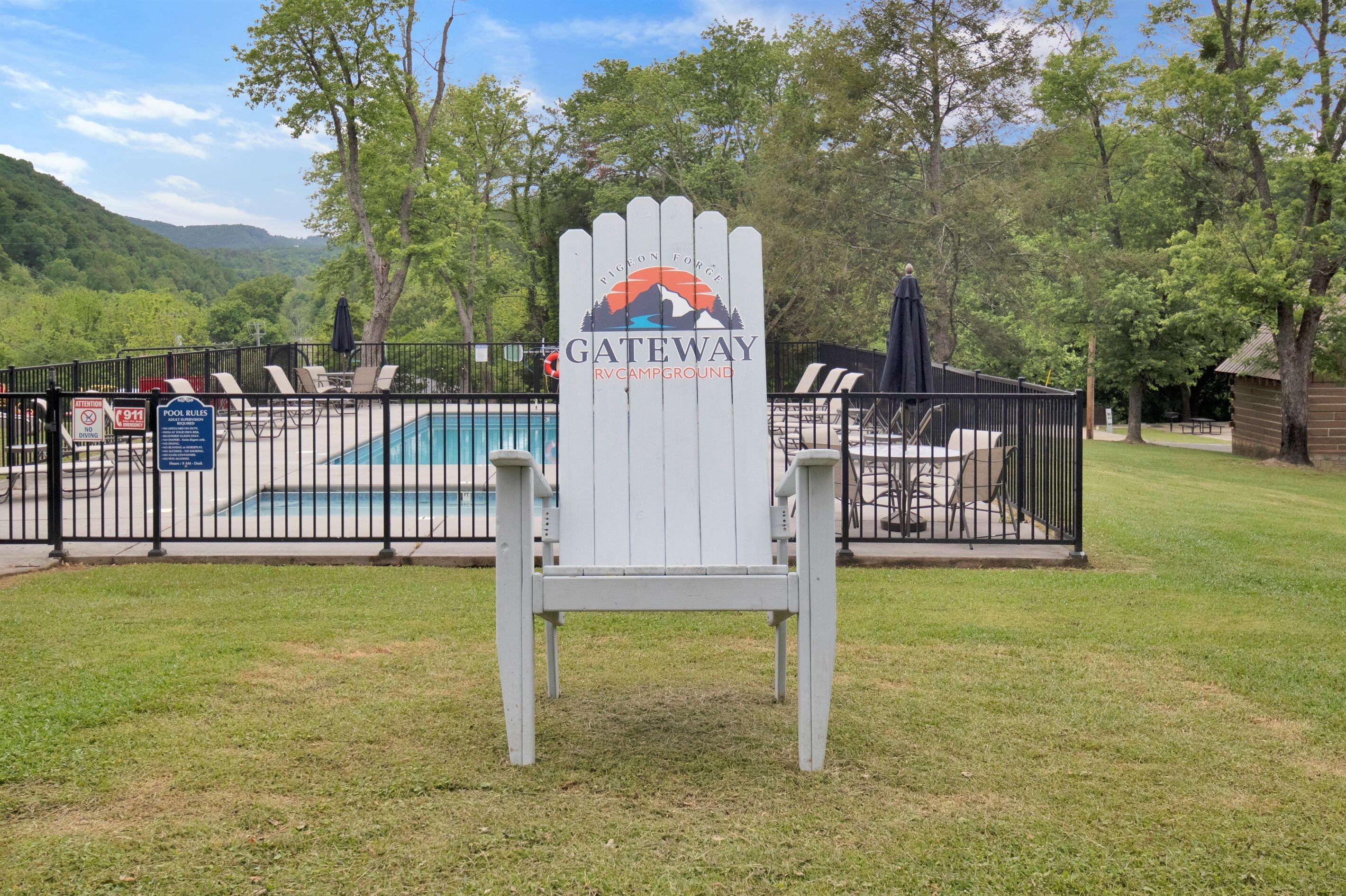 A white Adirondack chair with a Gateway RV Campground sign, a pool, and a fenced area with lounge chairs.