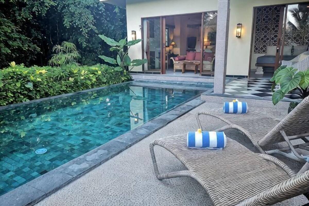 A swimming pool with lounge chairs and cushions, a covered seating area, and a view of a building with a glass door.