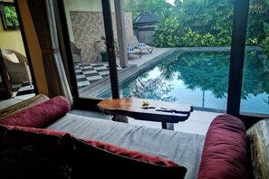 A room with a view of an infinity pool, a sofa, and a small table with a remote control. - Villa Nirvana (Bali)