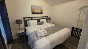 A neatly made bed with a gray and white striped blanket, two bedside lamps, a small bedside table, and a painting above the bed. - Pigeon Forge 2BR Cottage w/ King & Queen Beds (Pigeon Forge)
