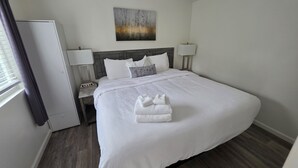A hotel room with a large bed, a bedside table with a lamp, a nightstand, a window with curtains, and a painting on the wall. - Pigeon Forge 2BR Cottage w/ King & Queen Beds (Pigeon Forge)