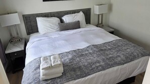 Ein ordentlich bezogenes Bett mit weißen Leinentüchern, einer grauen Tagesdecke und zwei gefalteten Handtüchern. Zwei Nachttischlampen, ein Nachttisch und ein an der Wand hängendes Bild sind ebenfalls zu sehen.