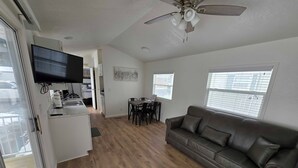Ein modernes Wohnzimmer mit grauem Ledersofa, einem Essbereich mit Tisch und Stühlen, einer Küche mit Fernseher und einem Deckenventilator.