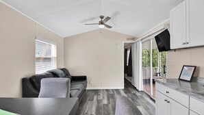 Un espacio habitable compacto con kitchenette, ventilador de techo, sofá, mesa de comedor y puerta corredera de vidrio que da a un área exterior.