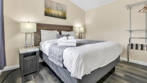 Habitación de hotel ordenada con una cama grande, dos mesitas de noche con lámparas, un perchero y un cuadro en la pared.
