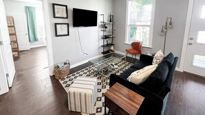 Un salon avec un canapé noir, un pouf à motifs et une table basse en verre. Un téléviseur est fixé au mur, il y a une bibliothèque et une fenêtre avec des stores.
