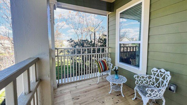 Une véranda en bois avec une balustrade blanche, deux chaises en fer forgé blanches, une petite table ronde ornée d’un vase, et une vue sur un jardin avec une clôture en bois et des arbres.