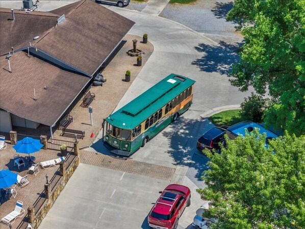 A green trolley bus parked in front of a building with a brick roof, a red car, and a blue car nearby.