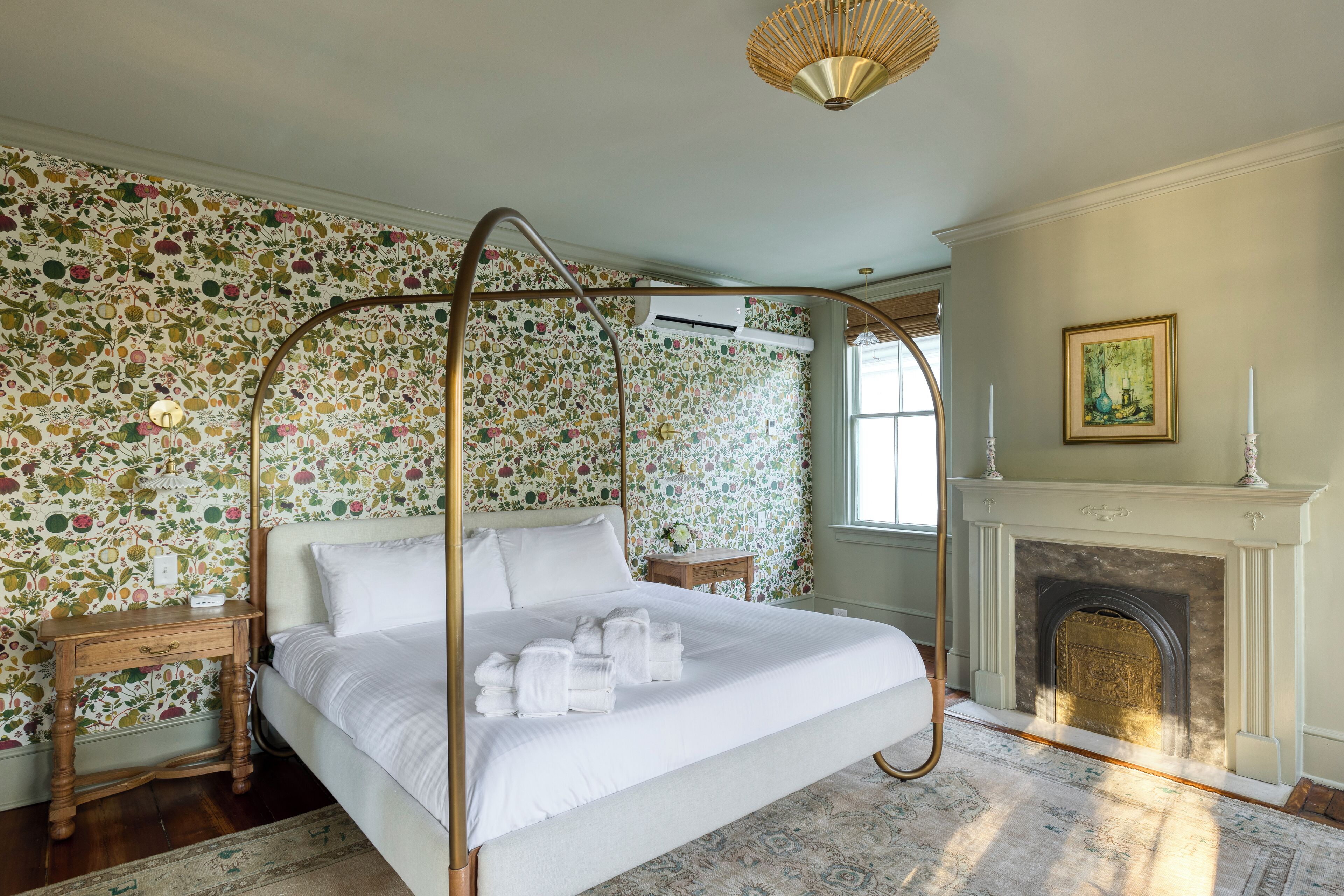 A four-poster bed with white linens, a wooden nightstand, a patterned rug, a fireplace, and floral wallpaper.