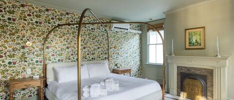 A four-poster bed with white linens, a wooden nightstand, a patterned rug, a fireplace, and floral wallpaper.