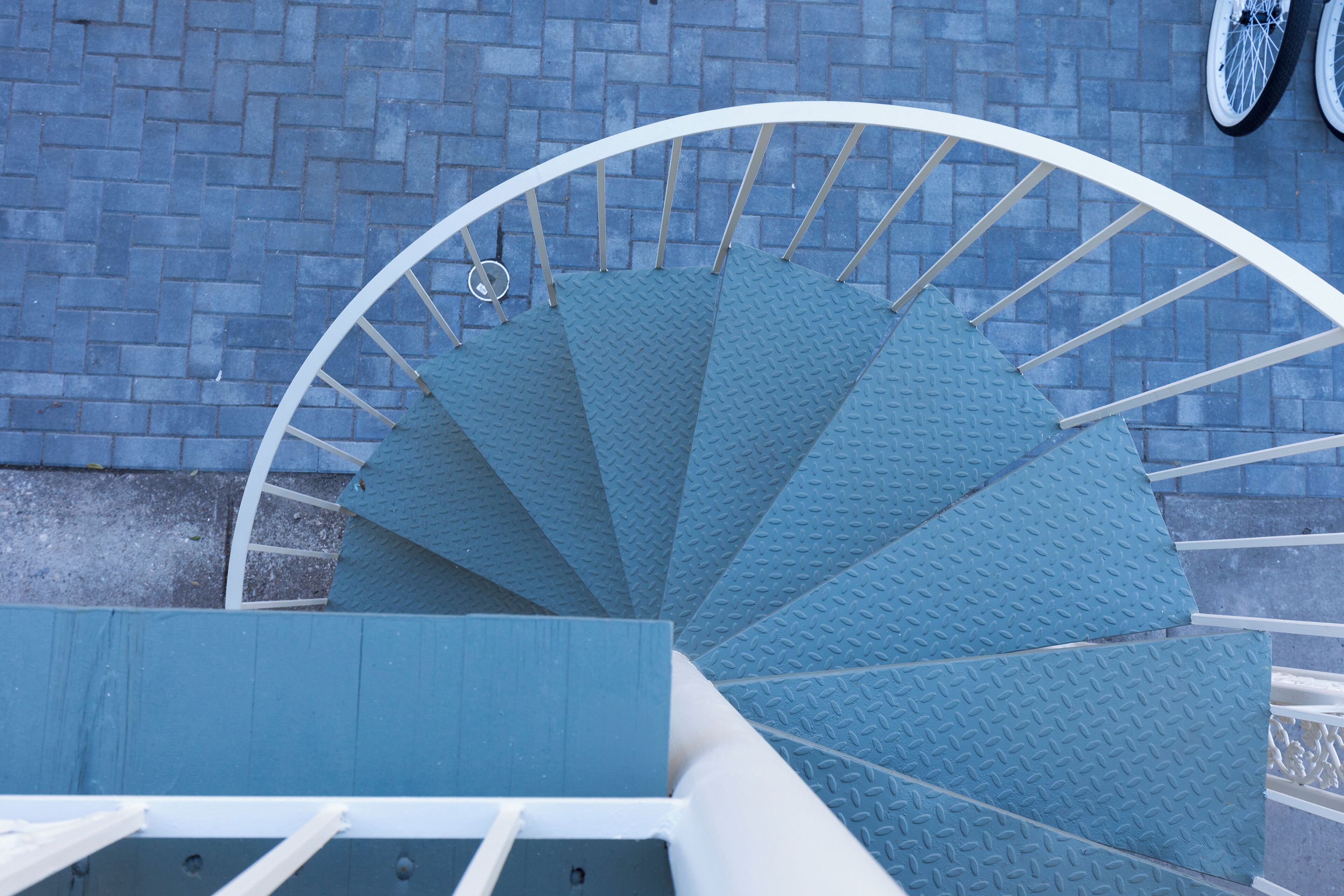 A spiral staircase with a blue textured handrail and steps, leading upwards against a brick wall background.
