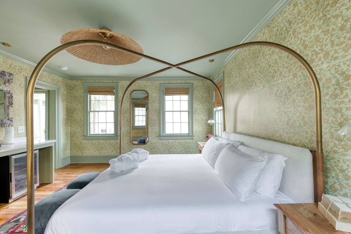 A spacious bedroom with a large bed, a desk, and a chair. There are two windows with curtains and a mirror on the wall.