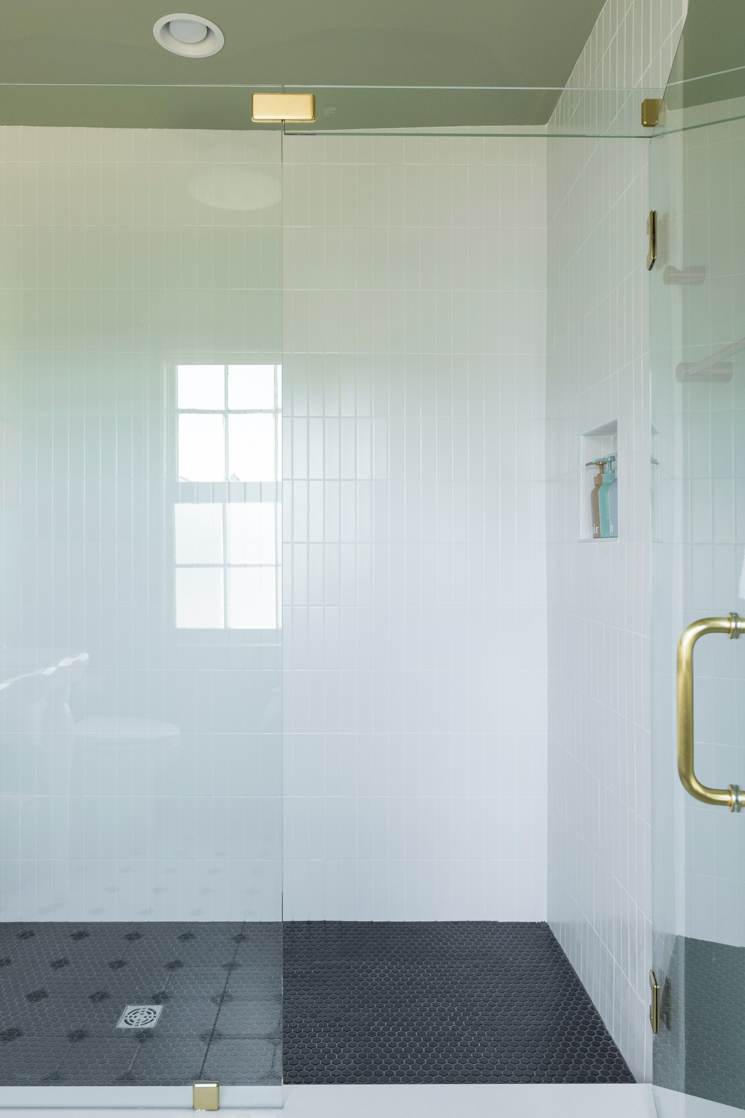 A glass-enclosed shower with a black textured floor, white tiled walls, and a small window.