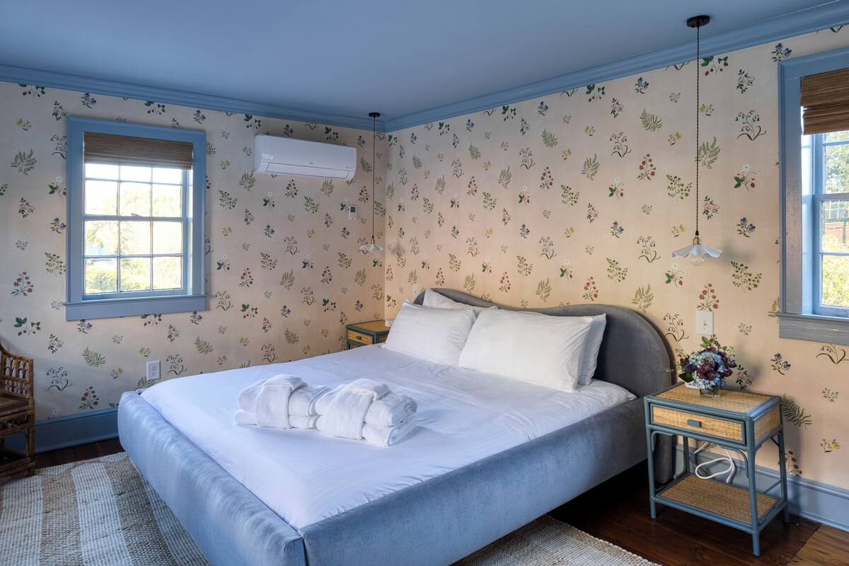 A neatly made bed with white linens and a towel, a small bedside table with a lamp, and a window with a view of greenery.