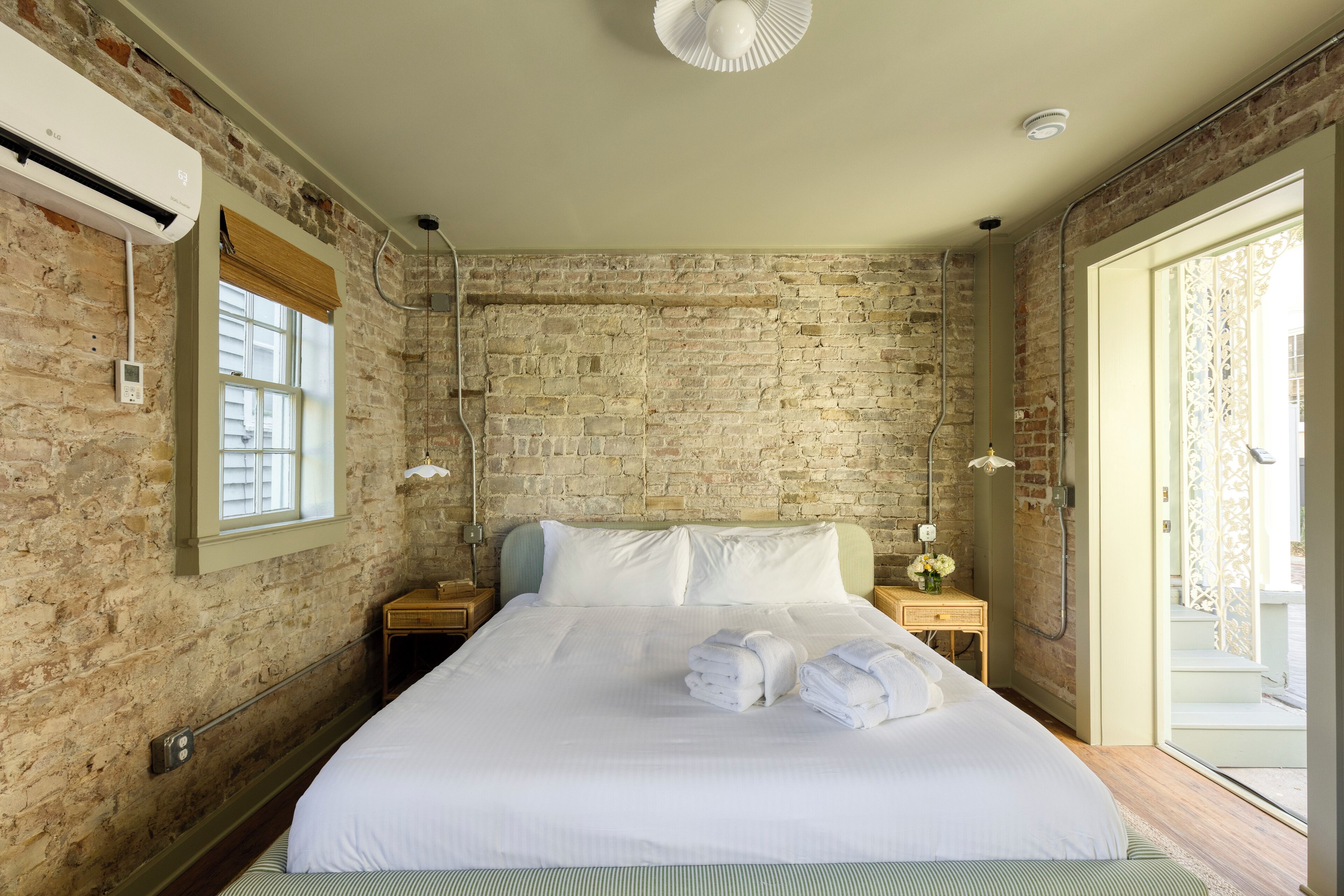 A bedroom with a brick wall, a large bed with white linens, a bedside table with a lamp, and a window with blinds.