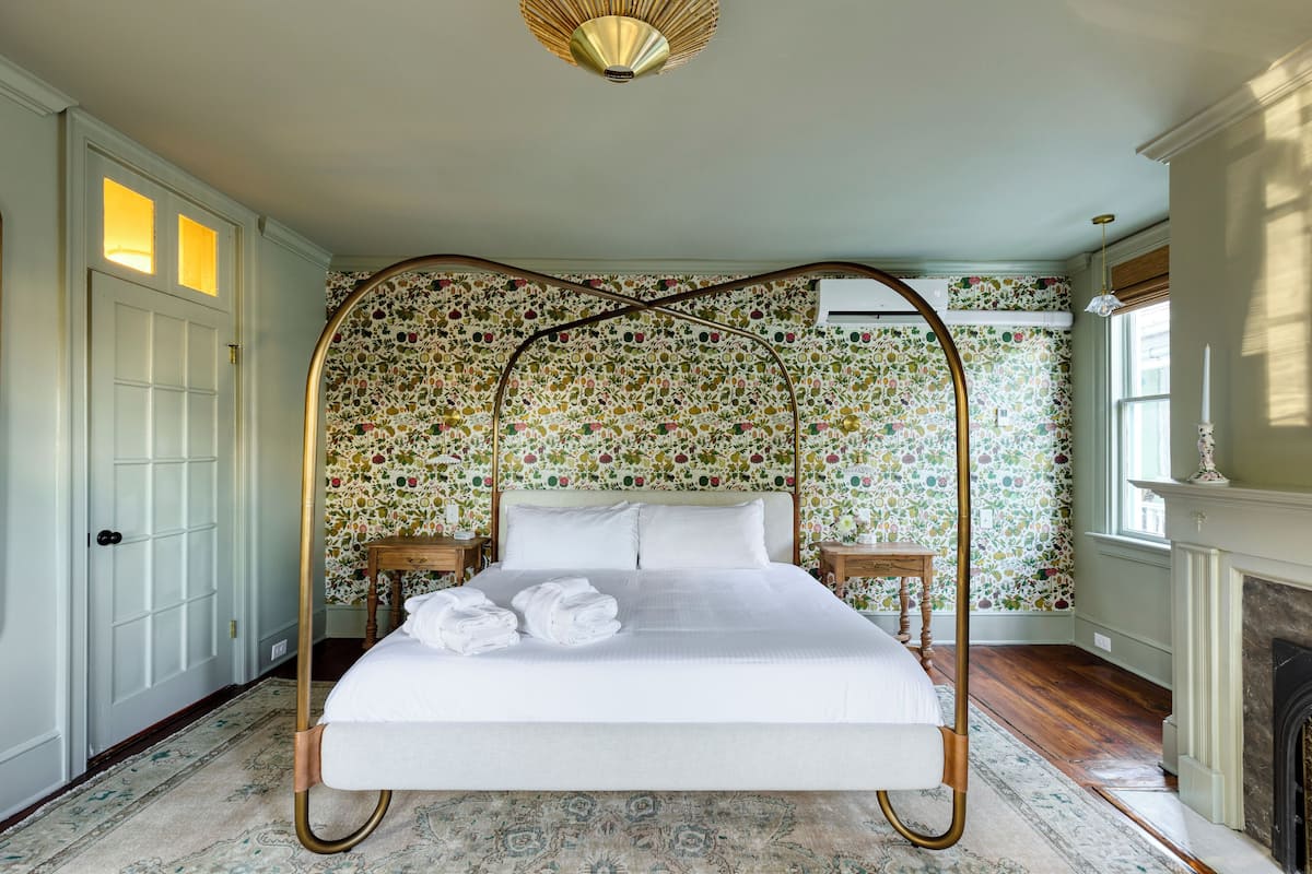 A four-poster bed with white linens, a floral wallpaper pattern, and a fireplace.