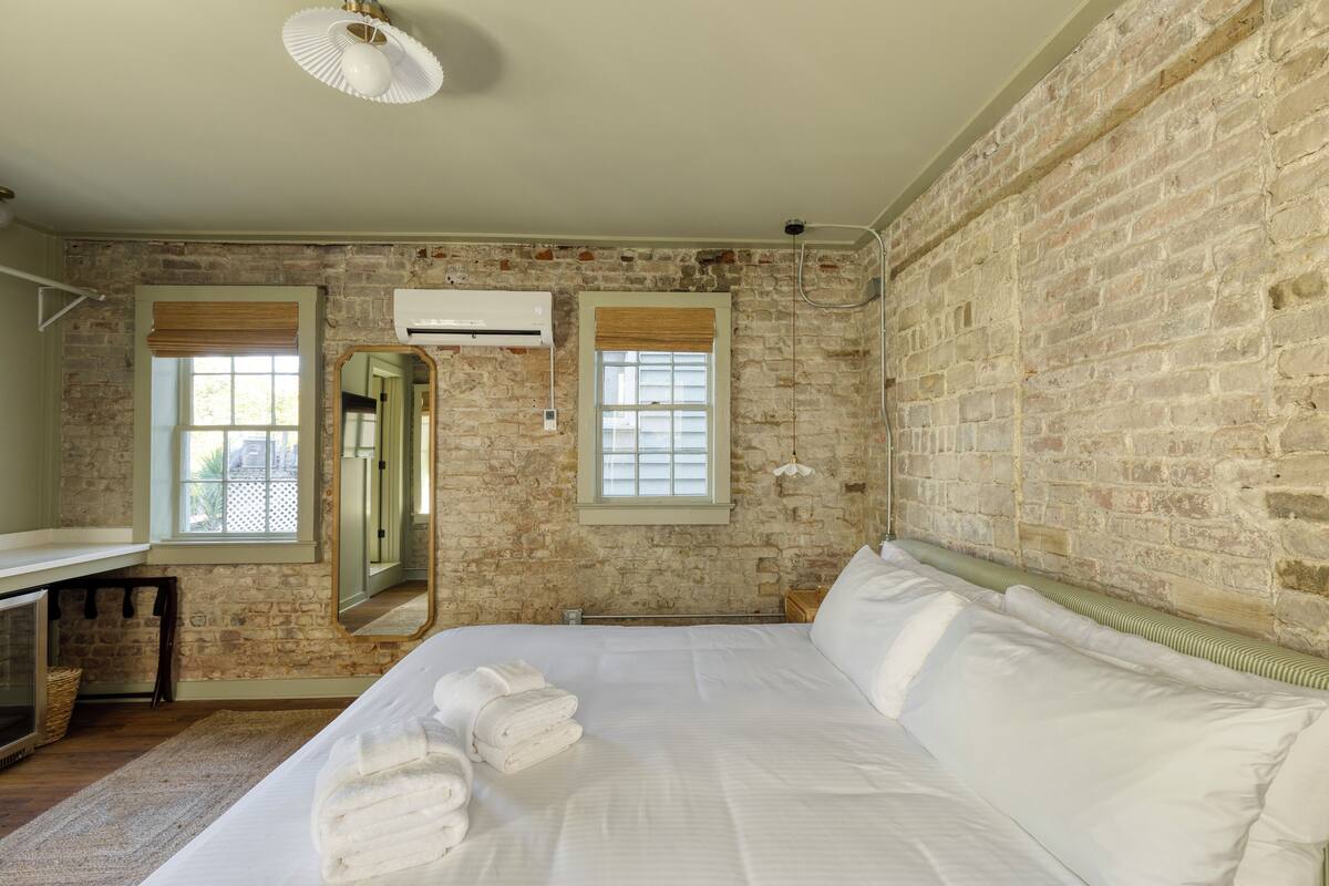 A bedroom with a brick wall, a large bed, a bedside table, and a window with blinds.