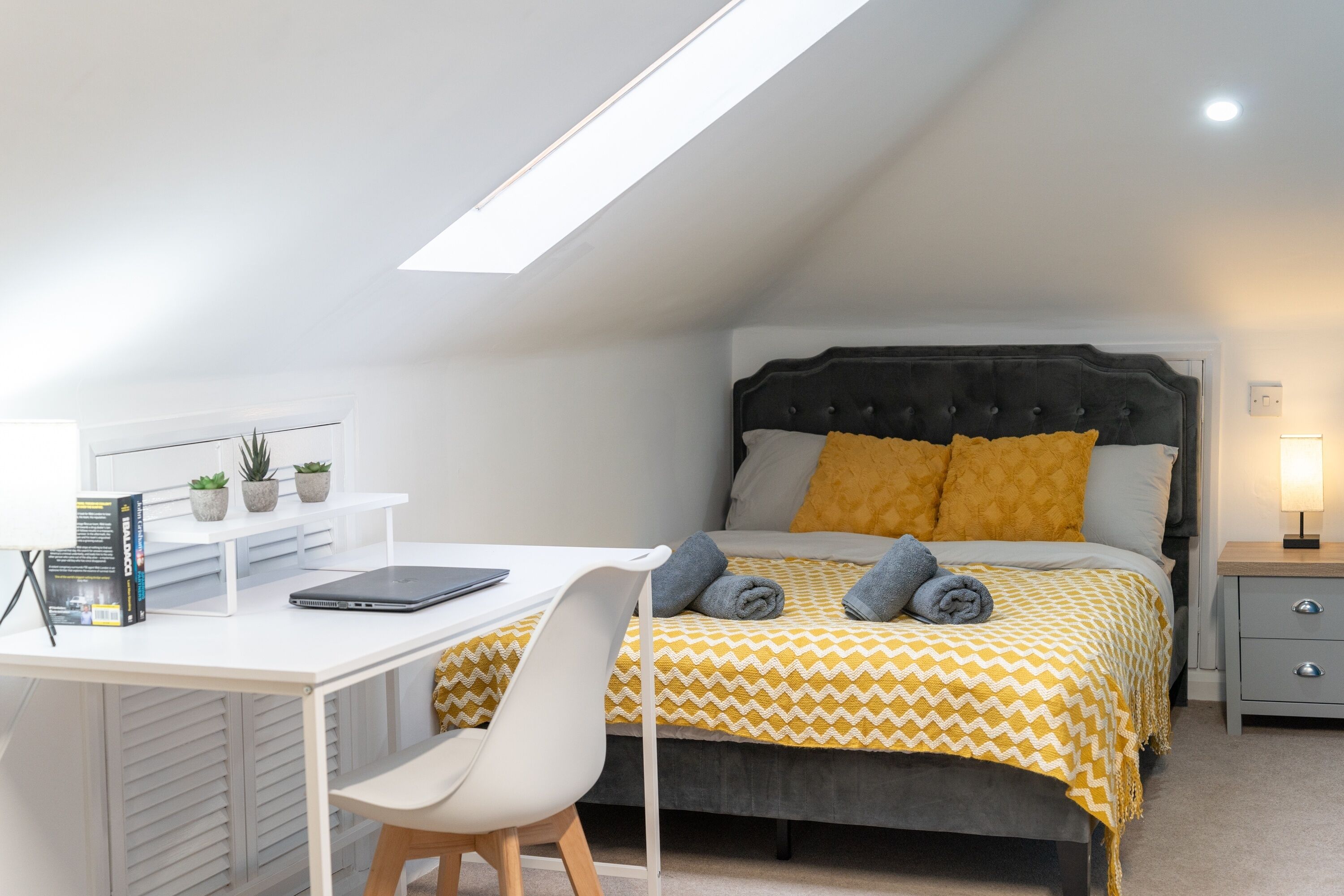 A bedroom with a bed, a desk, a chair, a bedside table, and a skylight.