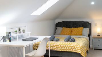 A bedroom with a bed, a desk, a chair, a bedside table, and a skylight.