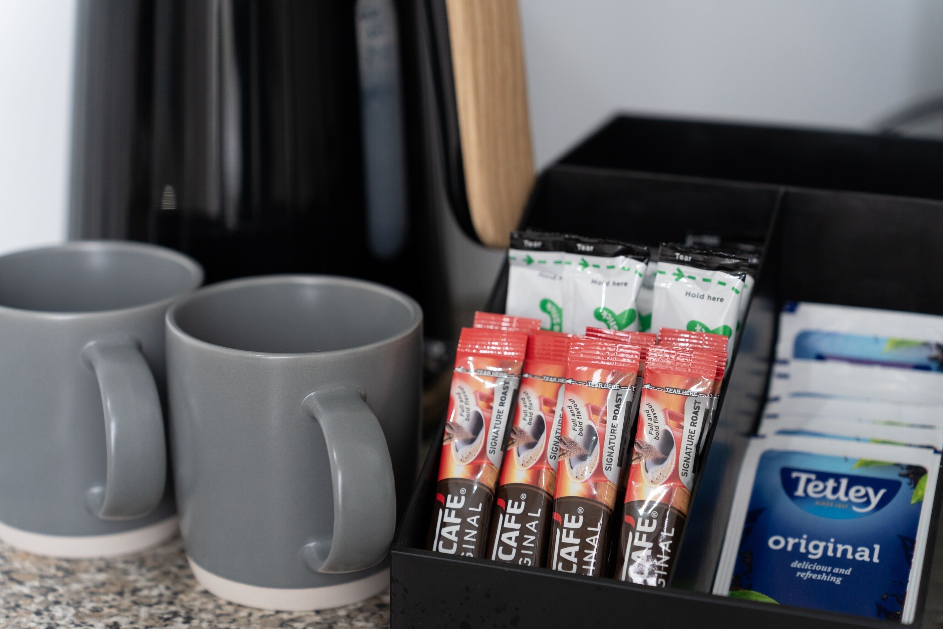 Eine Kaffeemaschine, zwei graue Tassen und eine Kapselkassette mit den Aufschriften 'Signature Roast' und 'Original' auf einer Arbeitsfläche.