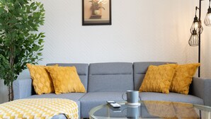 A modern living room with a grey sofa, yellow cushions, a glass coffee table, a remote control, and a framed picture on the wall.