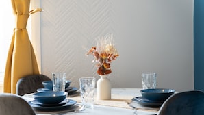 A dining table set with blue bowls, clear glasses, and a white vase with dried flowers.