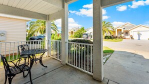 A covered outdoor patio with a table and chairs, a white fence, and a view of a residential street with houses and palm trees. - Hidden Palms! Next to Shipwreck Island! (Panama City Beach)