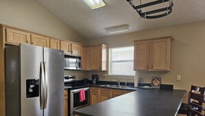 A modern kitchen with stainless steel appliances, wooden cabinets, a black countertop, and a window with blinds. - Hidden Palms! Next to Shipwreck Island! (Panama City Beach)