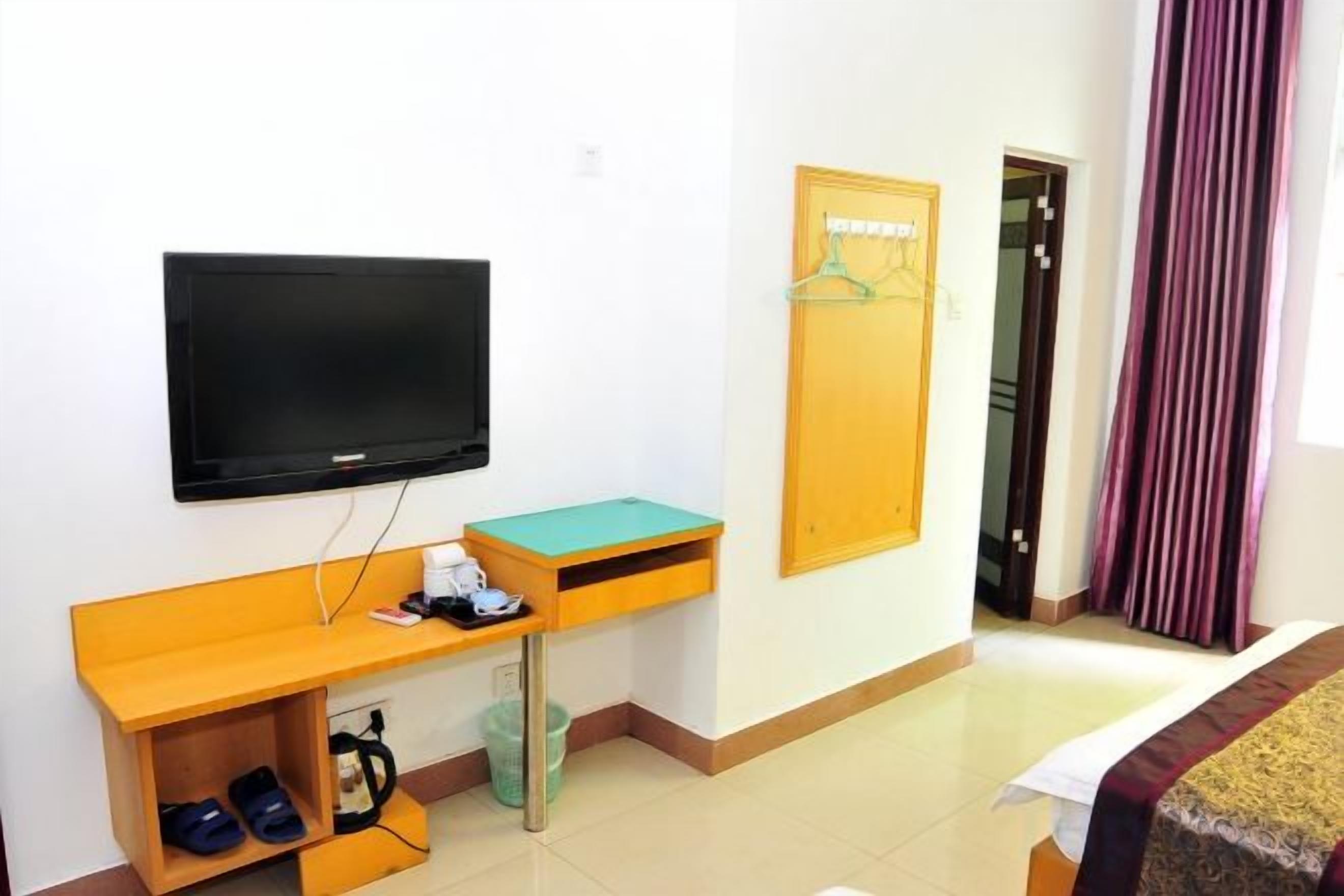 Ein Hotelzimmer mit an der Wand befestigtem Fernseher, einem Holztisch mit grüner Tischplatte, einem Bett mit gemustertem Überwurf und einer Tür mit violetten Vorhängen.